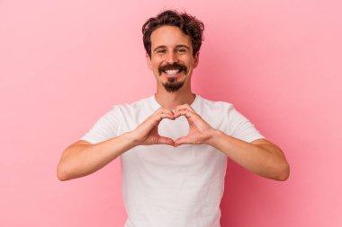 Pembe arka planda izole edilmiş genç beyaz adam gülümsüyor ve elleriyle kalp şeklini gösteriyor..