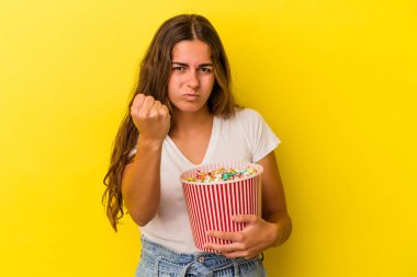 Sarı arka planda izole edilmiş patlamış mısır tutan genç beyaz kadın kameraya yumruk gösteriyor, agresif yüz ifadesi..