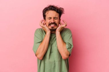 Pembe arka planda izole edilmiş genç beyaz adam kulaklarını elleriyle kapatıyor..