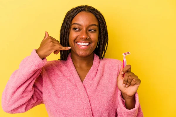 身穿浴衣的年轻的非洲裔美国女人拿着一把用黄色背景隔开的剃须刀片 手握着一个用手指打手机的手势 — 图库照片