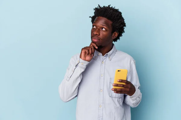 年轻的非洲裔美国人 手持蓝色背景的手机 侧视着对方 脸上带着怀疑和怀疑的表情 — 图库照片