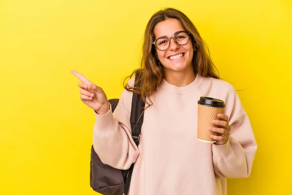 黄色の背景にコーヒーを閉じ込めた若い白人学生の女性は笑顔で脇を指差して 空白のスペースで何かを示す — ストック写真