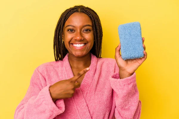 身穿浴衣的年轻的非洲裔美国女人拿着一块蓝色的海绵 背景是黄色的 面带微笑地把它放在一边 在空白的空间里展示着什么 — 图库照片
