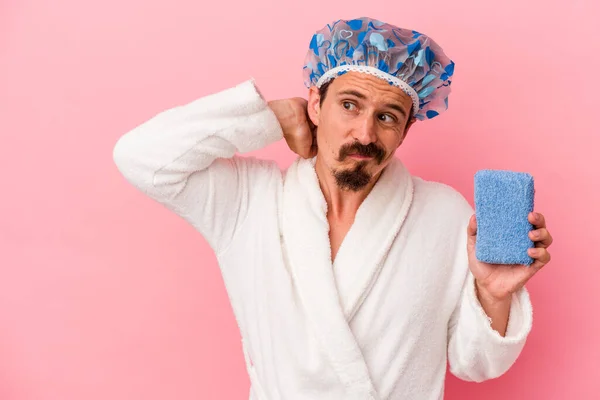 Young caucasian man going to the shower holding sponge isolated on pink background touching back of head, thinking and making a choice.