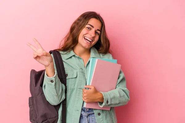 ピンクの背景に孤立した本を持つ若い白人学生の女性喜びと屈託のない指で平和のシンボルを示す — ストック写真
