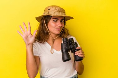 Sarı arka planda dürbün tutan genç beyaz kadın gülümseyerek beş numarayı parmaklarıyla gösteriyor..