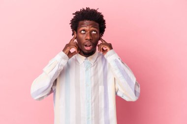 Genç Afrikalı Amerikalı bir adam pembe arka planda izole edilmiş parmaklarıyla kulaklarını kapatıyor, gürültülü bir ortam yüzünden stresli ve çaresiz..