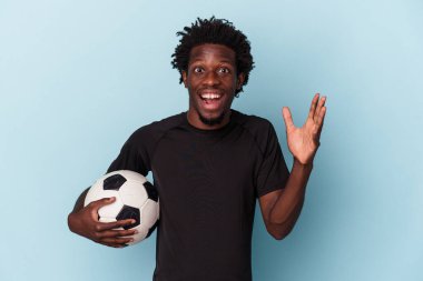 Amerikan futbolu oynayan genç Afro-Amerikan bir adam mavi arka planda izole edilmiş güzel bir sürpriz alıyor, heyecanlı ve el kaldırıyor..
