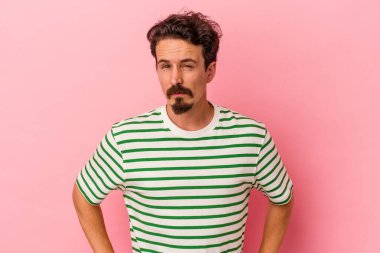 Pembe arka planda izole edilmiş genç beyaz adam hoşnutsuzca kaşlarını çatmış, kollarını kavuşturmuş..