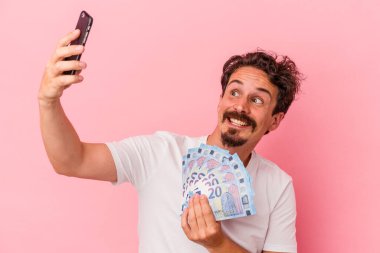 Elinde banknot tutan beyaz bir adam ve pembe arka planda izole edilmiş bir kumbara.