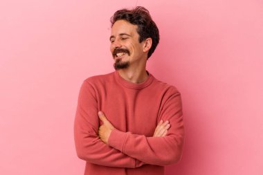 Pembe arka planda izole edilmiş genç beyaz adam kollarını kavuşturarak gülümsüyor..