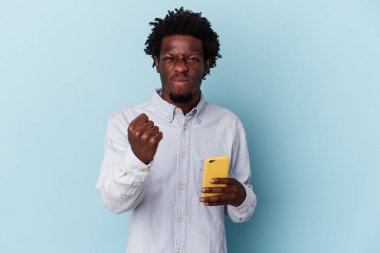 Elinde cep telefonu olan genç Afro-Amerikan bir adam mavi arka planda izole edilmiş kameraya yumruğunu gösteriyor, agresif bir yüz ifadesi var..