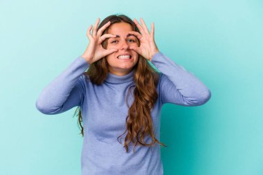 Mavi arka planda izole edilmiş genç beyaz kadın bir başarı fırsatı bulmak için gözlerini açık tutuyor..