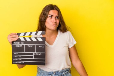 Sarı arka planda el çırpma tahtası tutan genç beyaz kadın kafası karışmış ve kendinden emin değil..