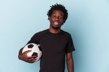 Futbol oynayan genç Afro-Amerikalı adam mavi arka planda izole edilmiş mutlu, güler yüzlü ve neşeli..