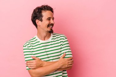 Pembe arka planda izole edilmiş genç beyaz adam kollarını kavuşturarak gülümsüyor..