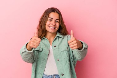 Pembe arka planda izole edilmiş genç beyaz kadın gülümsüyor ve başparmağını kaldırıyor.