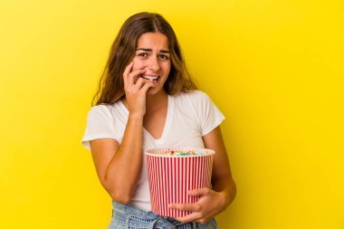 Elinde patlamış mısır tutan genç beyaz kadın sarı arka planda izole edilmiş tırnak yiyor, gergin ve çok endişeli..
