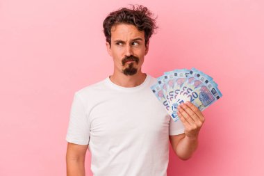 Pembe arka planda izole edilmiş banknotları tutan genç beyaz adam kafası karışmış, şüpheli ve güvensiz hissediyor..