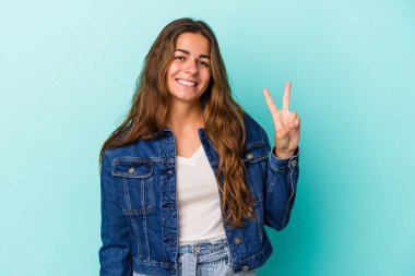 Mavi arka planda izole edilmiş genç beyaz kadın neşeli ve kaygısız parmakları olan bir barış sembolü gösteriyor..