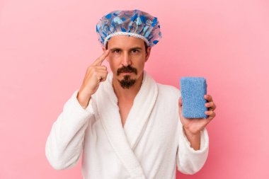 Duşa giren beyaz bir adam pembe arka planda izole edilmiş sünger tutuyor. Şakağı parmağıyla işaret ediyor, düşünüyor ve bir göreve odaklanıyor..