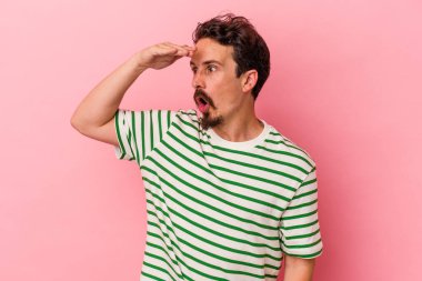 Pembe arka planda izole edilmiş genç beyaz adam uzaklara bakıyor ve elini alnına koyuyor..