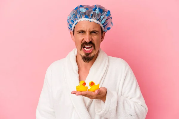 Young caucasian man going to the shower with rubber ducks isolated on pink background screaming very angry and aggressive.