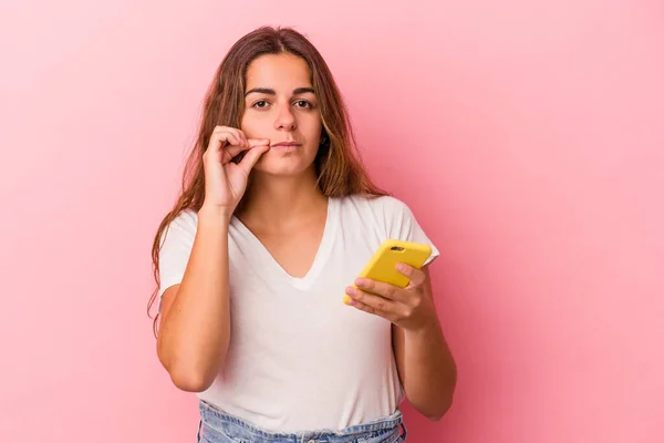 年轻的高加索女人用粉色背景隔离的手机 用手指捂着嘴唇保守秘密 — 图库照片