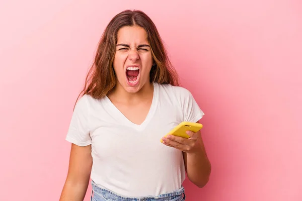 年轻的高加索女人用粉色背景隔离的手机尖叫着非常愤怒和咄咄逼人 — 图库照片