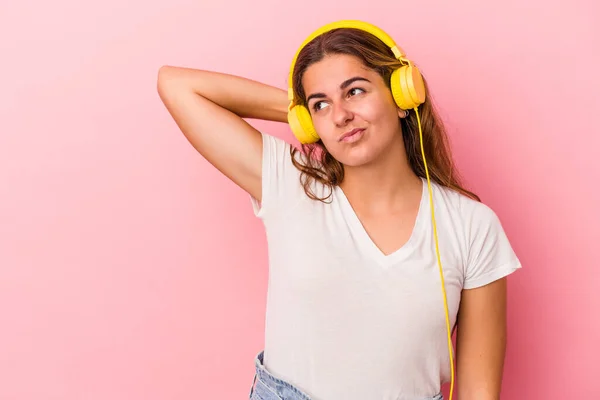 ピンクを背景に孤立した音楽を聴いている若い白人女性が頭に触れ 選択をする — ストック写真