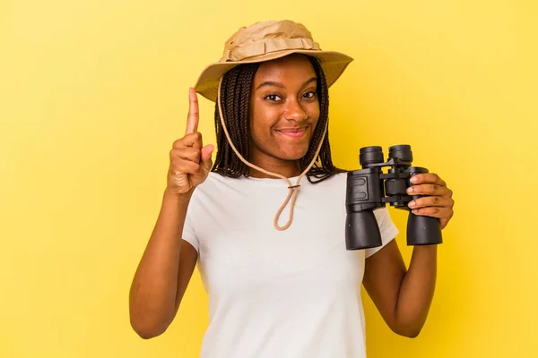 若いアフリカ系アメリカ人の探検家の女性は 指でナンバーワンを示す黄色の背景に隔離された双眼鏡を持って — ストック写真