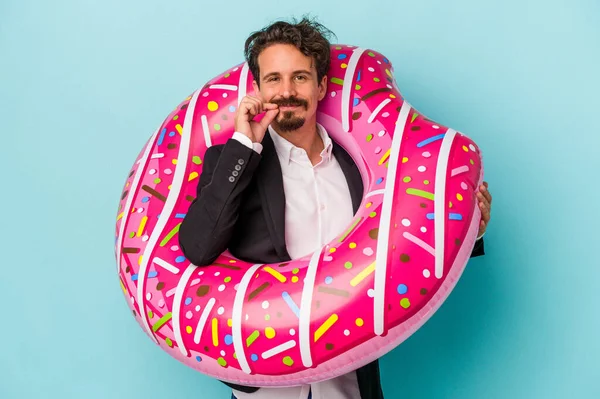 Young business man with inflatable donut isolated on blue background with fingers on lips keeping a secret.