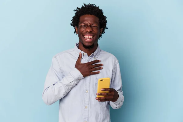 拿着蓝色背景的手机的年轻非洲裔美国人大声笑着把手放在胸前 — 图库照片