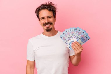 Elinde banknotlar tutan genç beyaz adam pembe arka planda izole edilmiş mutlu, güler yüzlü ve neşeli..