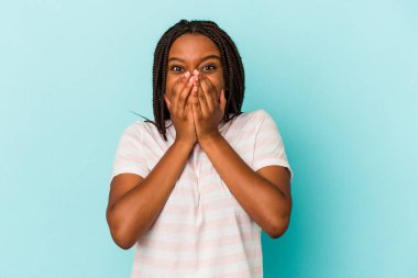 Genç Afrikalı Amerikalı bir kadın mavi arka planda izole edilmiş bir şekilde bir şeylere gülüyor, elleriyle ağzını kapatıyor..