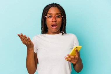 Elinde cep telefonu olan genç bir Afrikalı Amerikalı kadın mavi arka planda izole edilmiş. Şaşırmış ve şok olmuş..