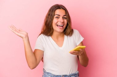 Pembe arka planda izole edilmiş cep telefonu kullanan beyaz bir kadın hoş bir sürprizle karşılaşıyor, heyecanlı ve el kaldırıyor..