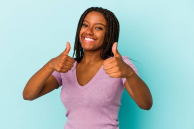 Genç Afrikalı Amerikalı bir kadın mavi arka planda izole edilmiş. Baş parmağını kaldırıyor, gülümsüyor ve kendinden emin..