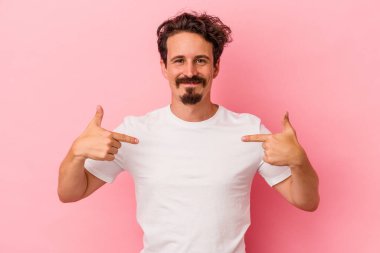 Pembe arka planda izole edilmiş genç beyaz bir adam gömlek fotokopi alanını elle gösteriyor, gururlu ve kendinden emin.