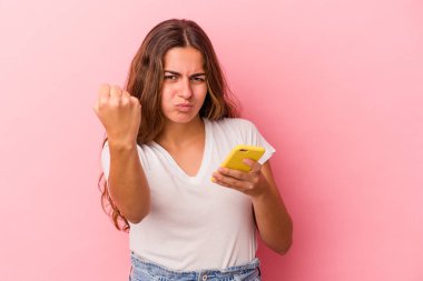 Pembe arka planda izole edilmiş cep telefonu kullanan genç beyaz kadın yumruğunu kameraya gösteriyor, agresif yüz ifadesi var..