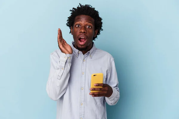拿着蓝色背景的手机的年轻的非洲裔美国人感到惊讶和震惊 — 图库照片