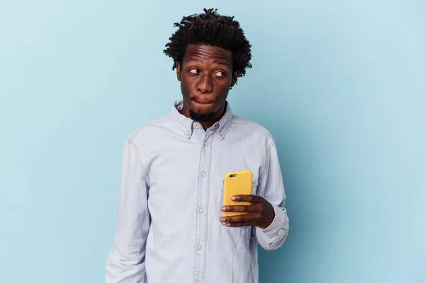 拿着蓝色背景的手机的年轻非洲裔美国人感到困惑 怀疑和不确定 — 图库照片