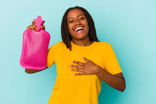 若いアフリカ系アメリカ人の女性が青い背景に隔離されたお湯の袋を持っています笑大声で胸に手を保つ — ストック写真