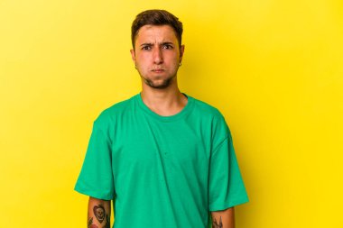 Sarı arka planda izole edilmiş dövmeleri olan beyaz genç bir adam yanakları patlatıyor, yorgun bir ifade takınıyor. Yüz ifadesi kavramı.