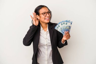 Elinde banknotlar tutan genç Latin kadın beyaz arka planda izole edilmiş kahve bir dedikoduyu dinlemeye çalışıyor..