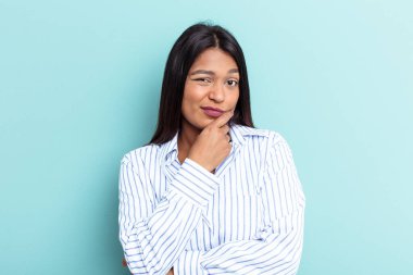 Genç Venezuelalı kadın mavi arka planda izole edilmiş şüpheli ve şüpheci bir ifadeyle yana bakıyor..