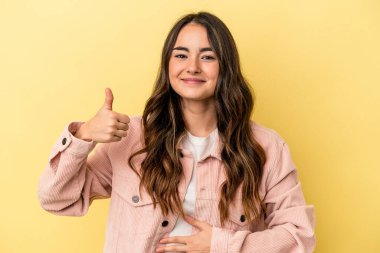 Sarı arka planda izole edilmiş genç beyaz kadın karnına dokunuyor, nazikçe gülümsüyor, yemek yiyor ve tatmin oluyor..