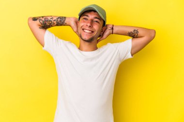 Sarı arka planda izole edilmiş, kollarını esneten, rahat bir pozisyonda dövmeleri olan genç beyaz bir adam..