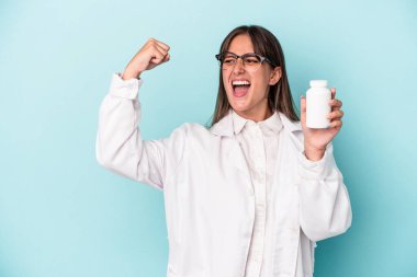 Mavi arka planda hapları tutan genç eczacı kadın zaferden sonra yumruğunu kaldırıyor, kazanan konsept..