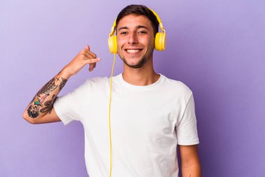 Mor arka planda izole edilmiş müzik dinleyen genç beyaz adam parmaklarıyla bir cep telefonu çağrısı hareketi gösteriyor..